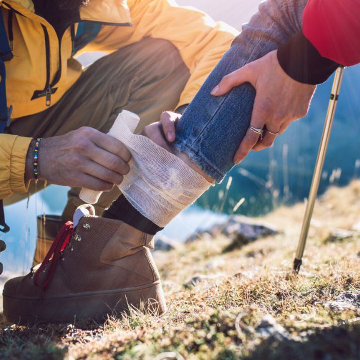 Injured hiker receiving first aid on mountain trail as companion wraps bandage around leg.
