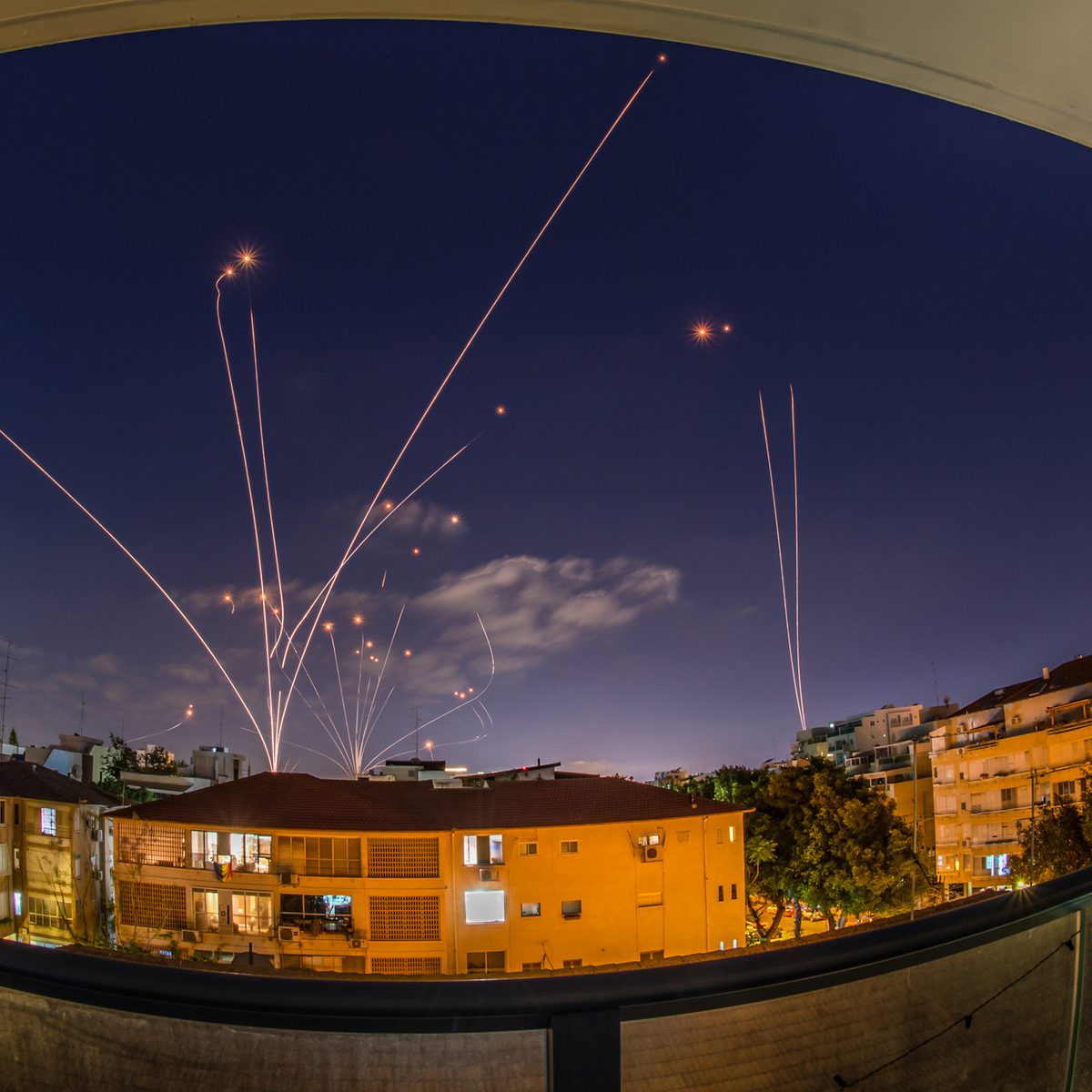 Israel’s Iron Dome air defence system intercepting rockets in the sky, with smoke trails and explosions visible above a cityscape at dusk.